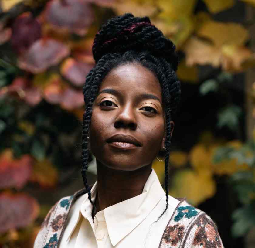 A woman with plaited hair styled in a bun stands outdoors, wearing a white collared shirt and patterned jumper. She faces the camera with a calm expression, as if ready for a coffee-break interview; blurred autumn leaves frame Evie Asio in the background.