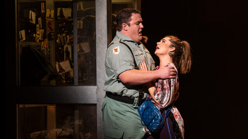 John Findon, Ava Dodd, ENO’s Carmen 2025 © Ellie Kurttz A man in a uniform and a woman with a blue handbag embrace passionately beside a vintage phone box labelled Teléfono, under dramatic stage lighting.