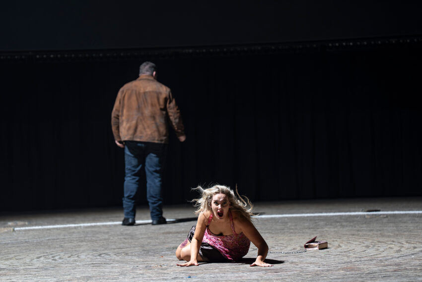 John Findon, Niamh O'Sullivan, ENO’s Carmen 2025 © Ellie Kurttz A woman in a pink dress is on the floor, looking distressed, whilst a man in a brown jacket stands with his back to her on a dimly lit stage. A pair of high-heeled shoes lies nearby.