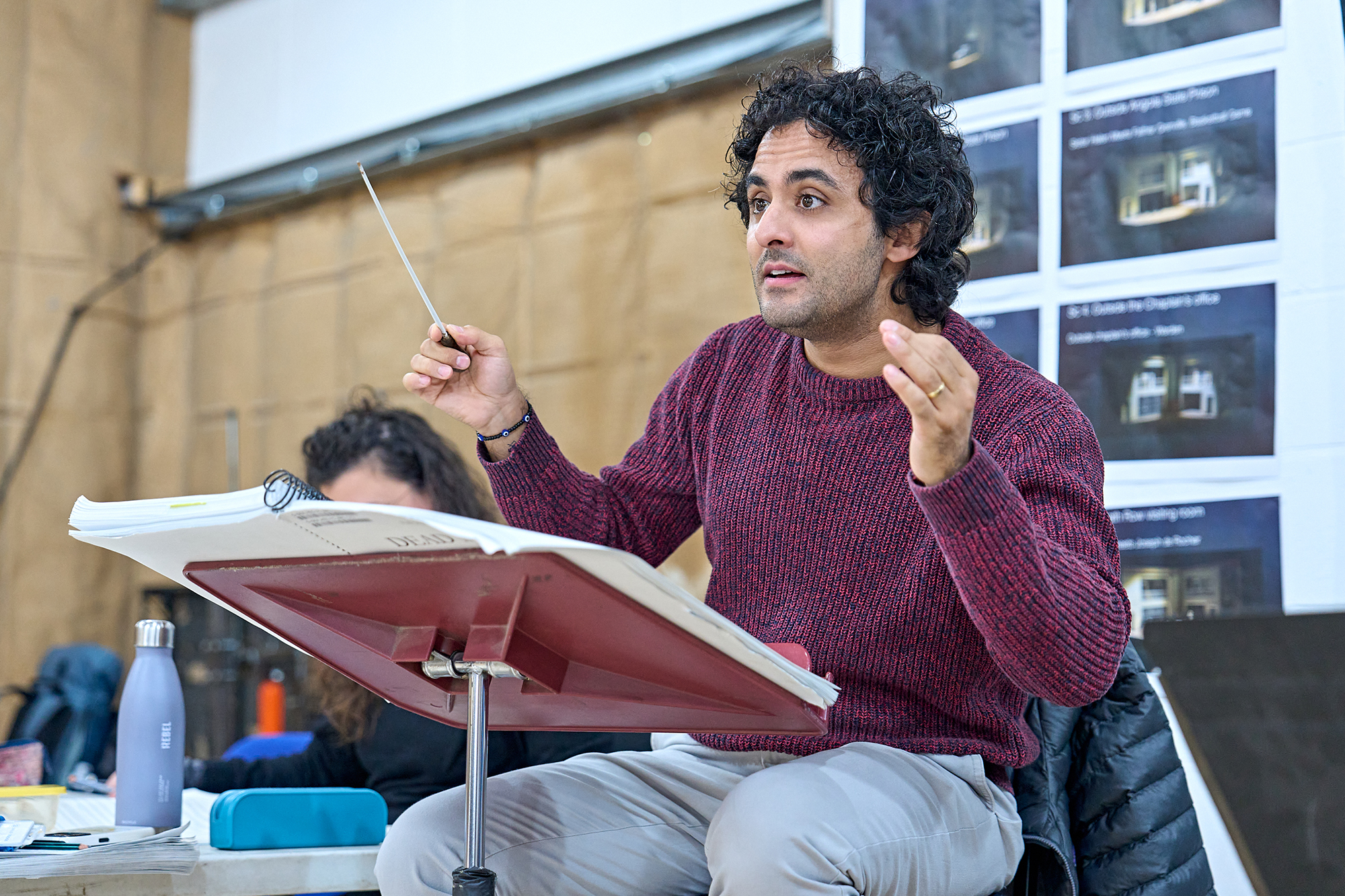 Kerem Hasan, a man with curly hair wearing a maroon jumper sits at a music stand, holding a conductor’s baton and gesturing with his other hand, appearing to lead or explain something. Sheet music and notes are visible in front of him.
