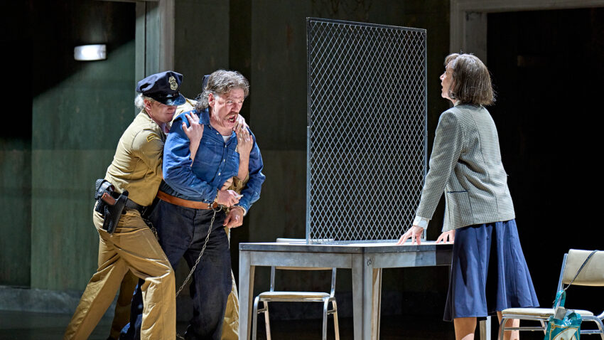 A police officer restrains a distressed man beside a table with a metal divider, while a woman in a skirt and blazer leans forward on the opposite side, appearing tense and engaged in conversation.