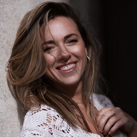 A woman with long, light brown hair smiles whilst leaning against a light-coloured wall. She is wearing a white crocheted top and gold hoop earrings, and sunlight highlights her face and hair.