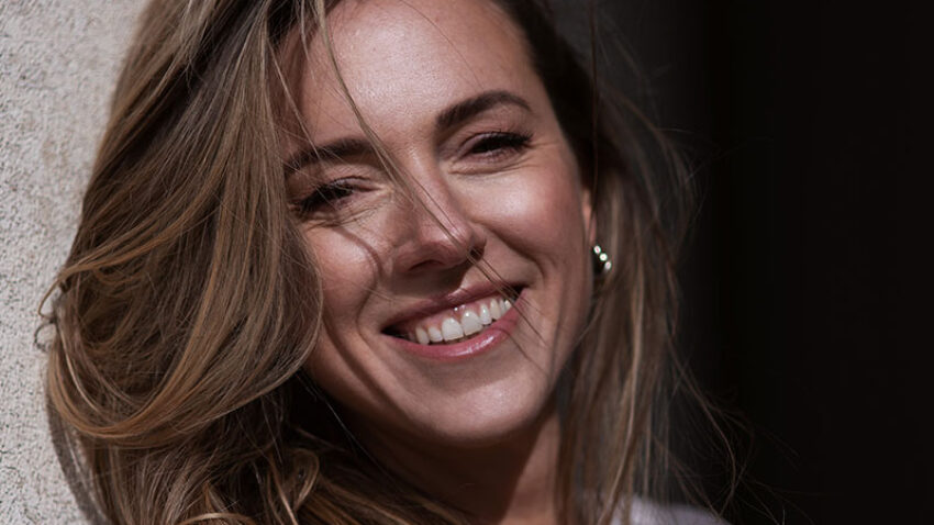 A woman with long, light brown hair smiles whilst leaning against a light-coloured wall. She is wearing a white crocheted top and gold hoop earrings, and sunlight highlights her face and hair.