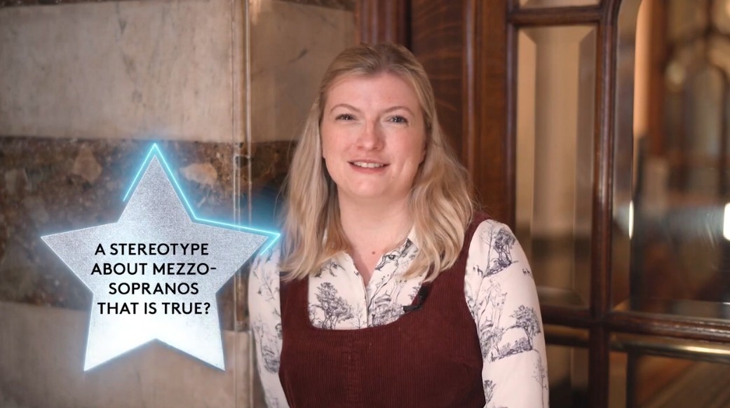 A smiling woman with long blonde hair, wearing a patterned blouse and maroon sleeveless dress, sits indoors. Beside her, a glowing star graphic reads: A stereotype about mezzo-sopranos that is true? ENO Harewood Artists support.