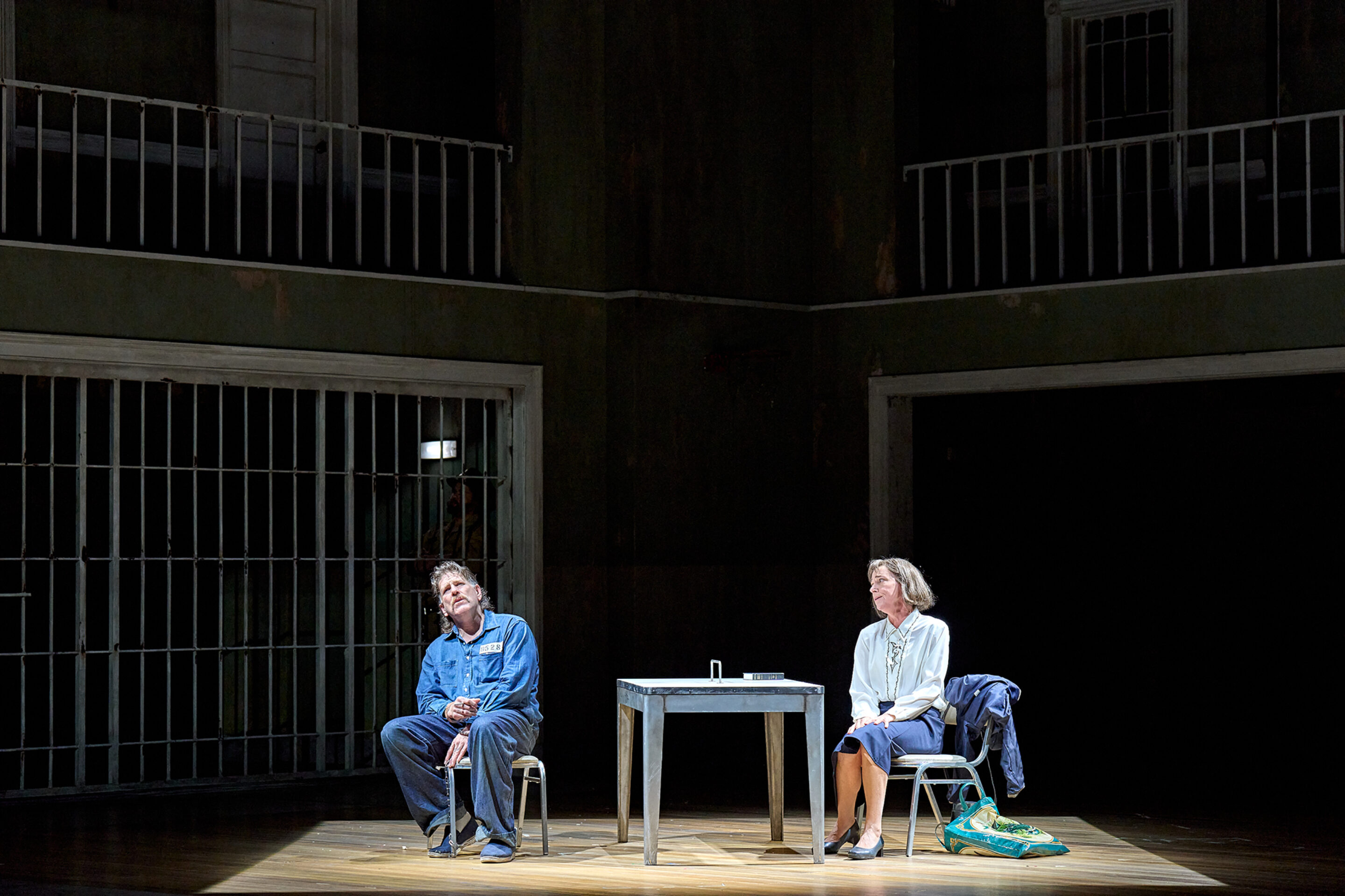 A man in blue sits slouched on a stool and a woman in a white blouse and blue skirt sits upright on a chair at a small table on a dimly lit stage with barred railings in the background.