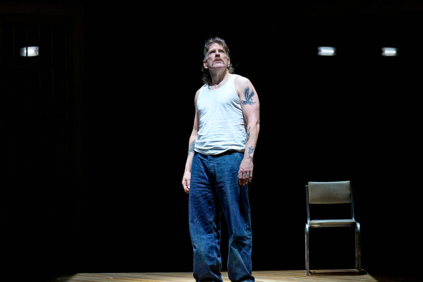 A man in a white vest and blue trousers stands barefoot on a wooden stage, looking up with a serious expression. A metal chair is behind him, and the background is dark with a few small lights visible.