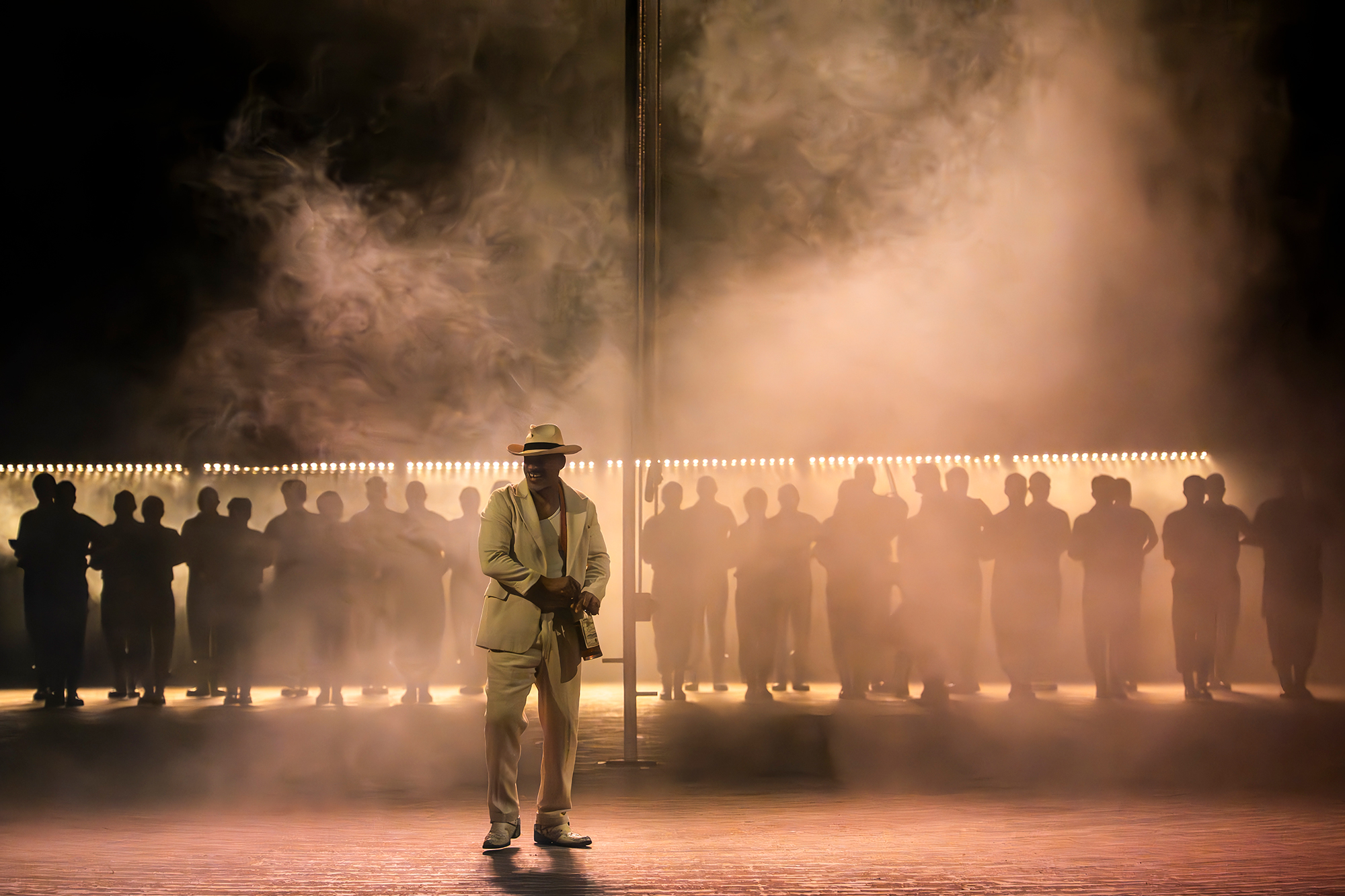 The Cast of ENO’s Carmen 2025 © Ellie Kurttz A person in a white suit and hat stands on stage in dramatic lighting and fog, with a row of people in shadow behind them, creating a mysterious and theatrical atmosphere.
