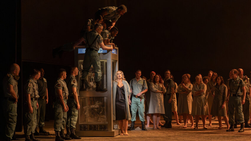The Cast of ENO’s Carmen 2025 © Ellie Kurttz A woman in a black dress sings on stage, surrounded by men in military uniforms and women in dresses. Some men are standing on top of a glass structure, while others watch intently under dramatic lighting.