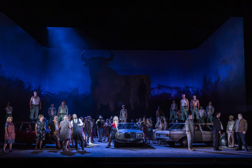 The Cast of ENO’s Carmen 2025 © Ellie Kurttz A dramatic stage scene features a large cast in modern dress, standing around cars under blue lighting. A silhouette of a bull is projected on the backdrop, and some performers stand on top of the cars, creating a tense atmosphere.