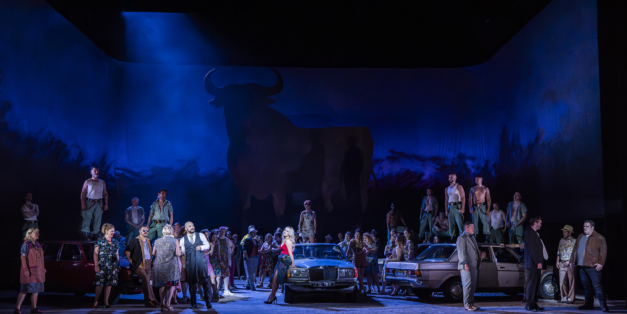 The Cast of ENO’s Carmen 2025 © Ellie Kurttz A stage scene features a large group of people around two vintage cars, with some standing on the vehicles. The background shows a dramatic blue-lit wall with a black silhouette of a bull.