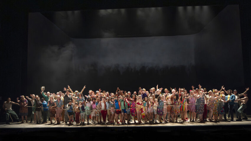 The Cast of ENO’s Carmen 2025 © Ellie Kurttz A large group of people stand together on a stage, many in colourful clothing, raising their arms and pointing upward. The background is dark with dramatic lighting above, suggesting a theatre performance or final curtain call.