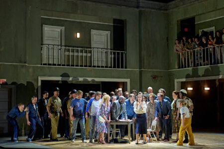 A large group of people gathers closely around a table in the centre of a dimly lit, two-storey room with green walls, while more people look on from a balcony above. The scene is tense and dramatic.