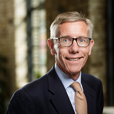 A smiling middle-aged man with short fair hair and glasses, wearing a navy suit, light blue shirt, and orange patterned tie, stands indoors with a blurred background of windows and brick walls.