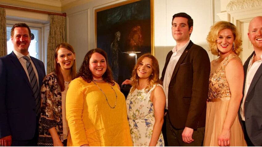 Seven adults, including Harewood Artists, dressed in formal and semi-formal attire, stand side by side smiling at the camera in a warmly lit, elegant room with paintings and decorative curtains in the background.