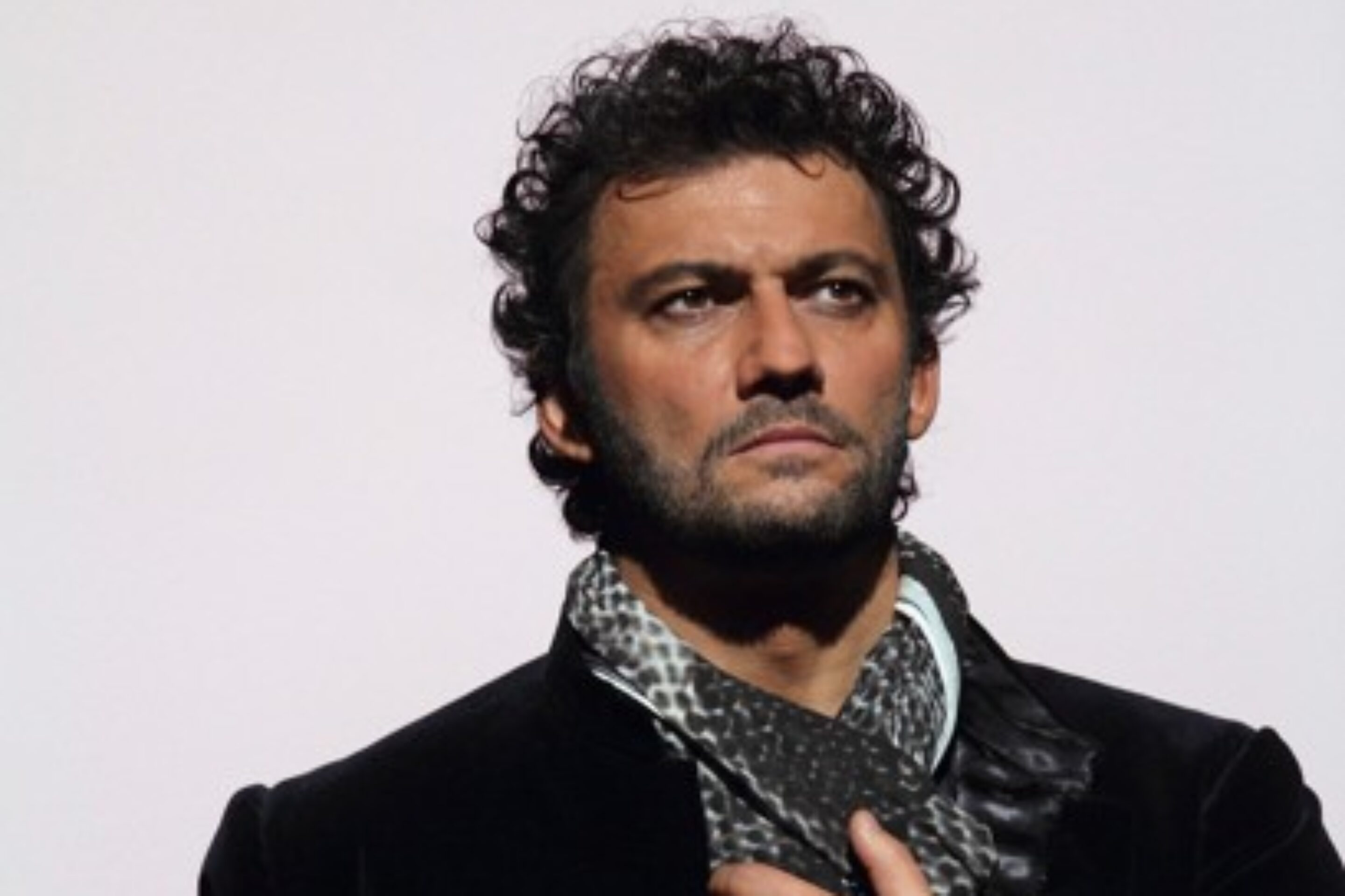 Image of the tenor singer Jonas Kaufmann - A man with curly dark hair and a trimmed beard, wearing a dark jacket and a patterned scarf, stands against a plain light background, looking serious.