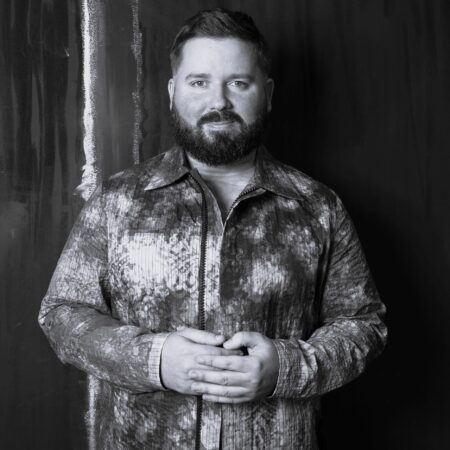 A bearded man with short hair stands against a dark background, wearing a patterned, button-up shirt. Kip Williams faces the camera, hands loosely clasped and serious expression. The photo is in striking black and white.