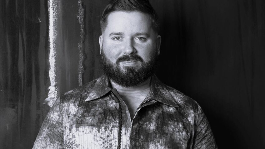 A bearded man with short hair stands against a dark background, wearing a patterned, button-up shirt. Kip Williams faces the camera, hands loosely clasped and serious expression. The photo is in striking black and white.