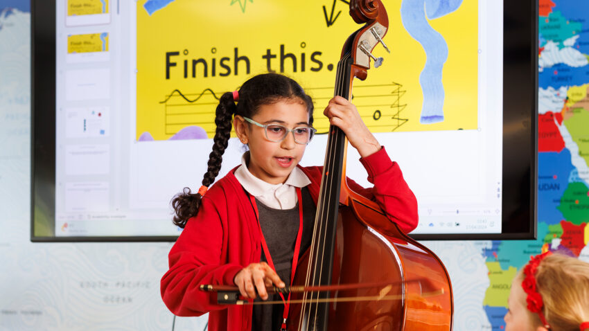 A young girl in a red jumper plays a double bass with a bow in a classroom. Behind her, a screen displays a yellow ENO Curriculum slide reading “Finish this.” and musical notes. Another child watches in the foreground.