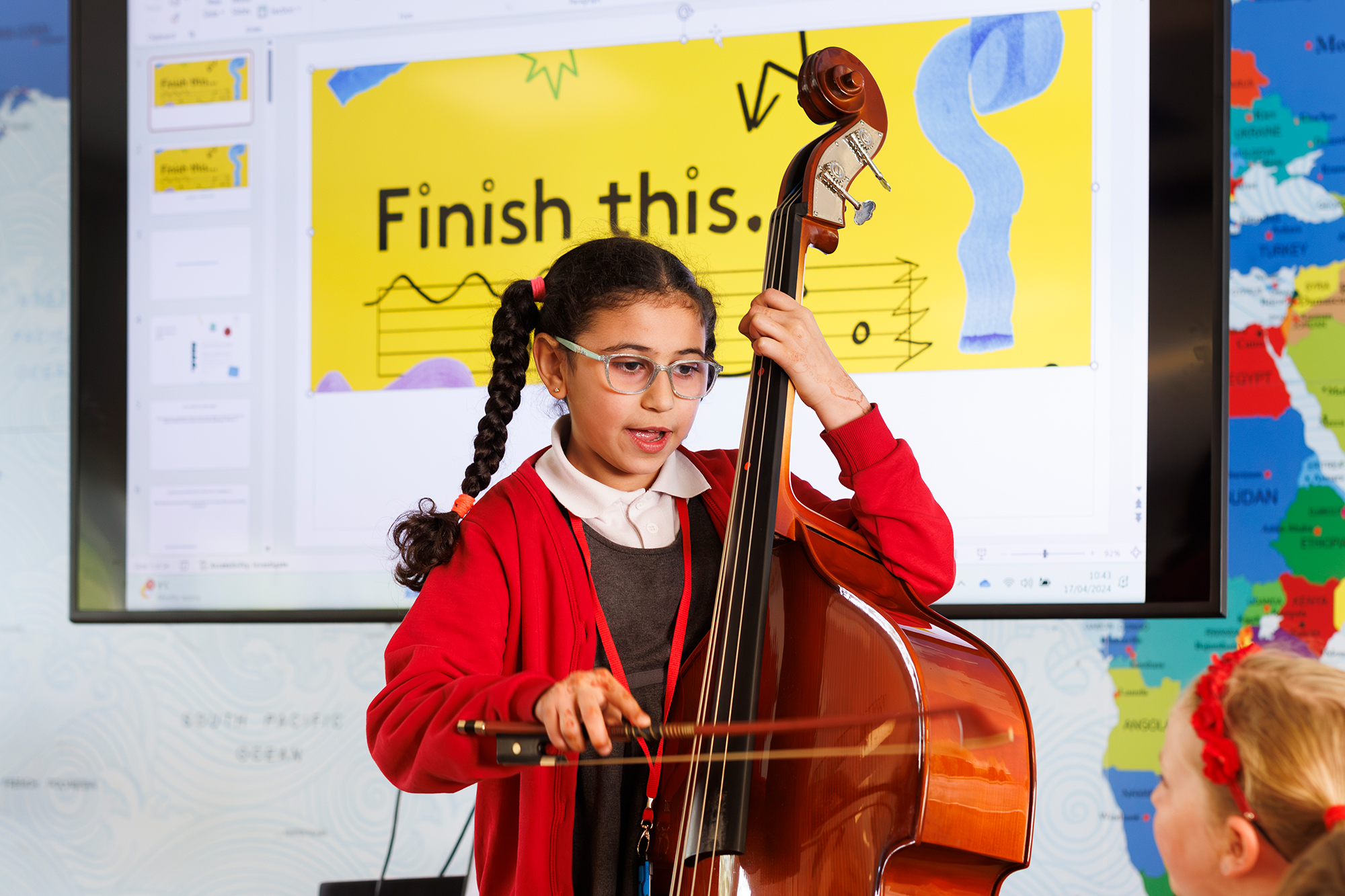 A young girl in a red jumper plays a double bass with a bow in a classroom. Behind her, a screen displays a yellow ENO Curriculum slide reading “Finish this.” and musical notes. Another child watches in the foreground.