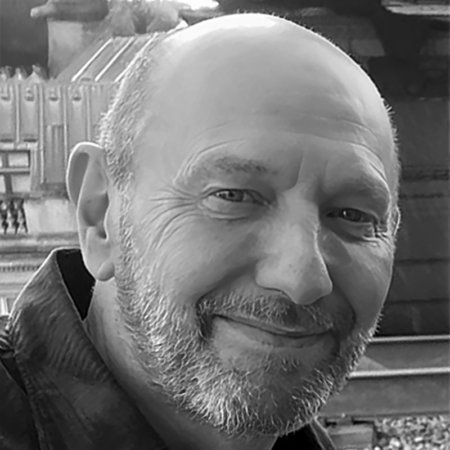 Adrian Plaut, A man with a beard and a bald head smiles slightly in this black and white photo, taken outdoors in front of a building with ornate architectural details.