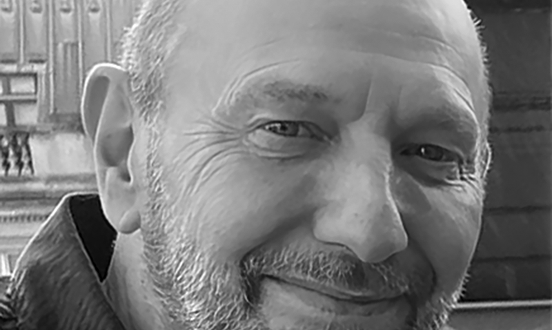 Adrian Plaut, A man with a beard and a bald head smiles slightly in this black and white photo, taken outdoors in front of a building with ornate architectural details.