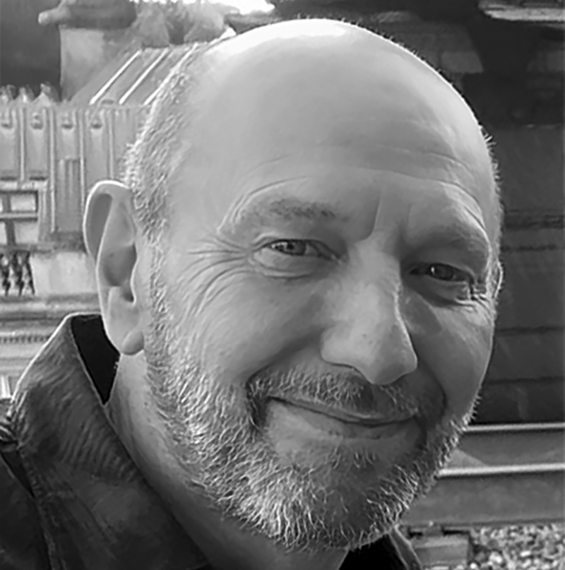 Adrian Plaut, A man with a beard and a bald head smiles slightly in this black and white photo, taken outdoors in front of a building with ornate architectural details.