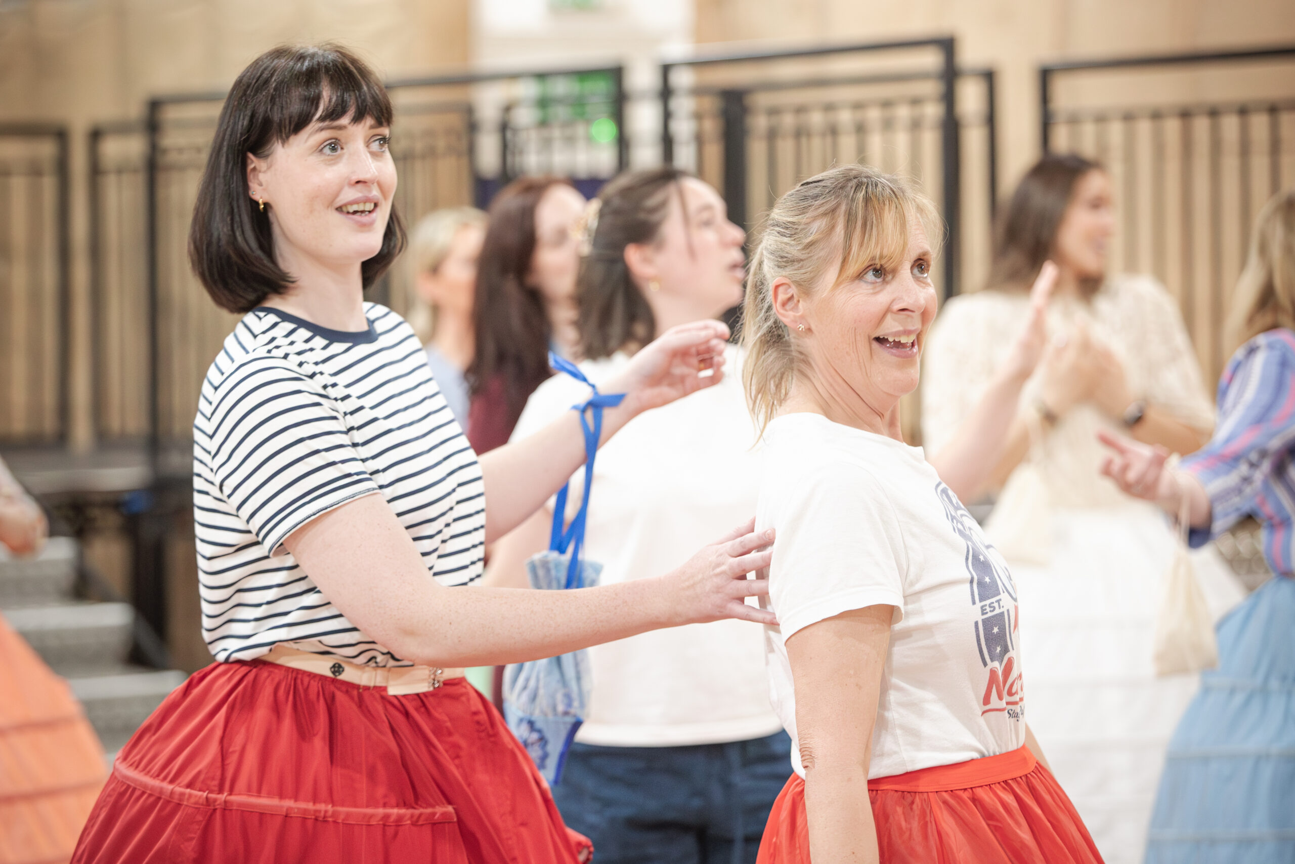 A group of women smiling and standing in a queue indoors, wearing brightly coloured skirts and casual tops—two in the foreground appear cheerful and engaged, as if awaiting an interview with Mel Giedroyc about their lively HMS Pinafore-inspired activity.
