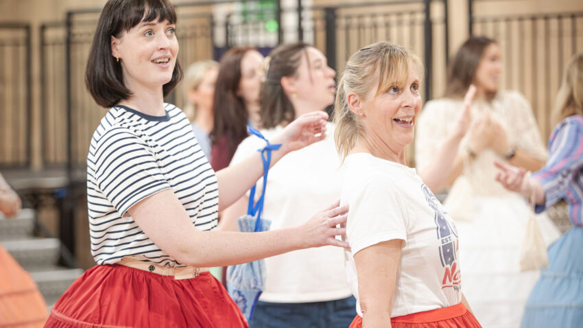 A group of women smile and laugh together indoors. Bethan Langford and Mel Giedroyc in front wear vintage-style skirts and are engaged in an interactive activity, while others chat and stand in the background.