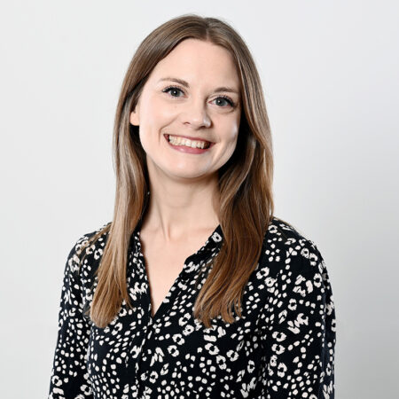 Caroline Julian, with straight, light brown hair, is smiling at the camera. She is wearing a black blouse with a white spotted pattern and stands against a plain, light grey background.