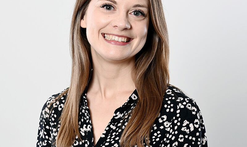 Caroline Julian, with straight, light brown hair, is smiling at the camera. She is wearing a black blouse with a white spotted pattern and stands against a plain, light grey background.