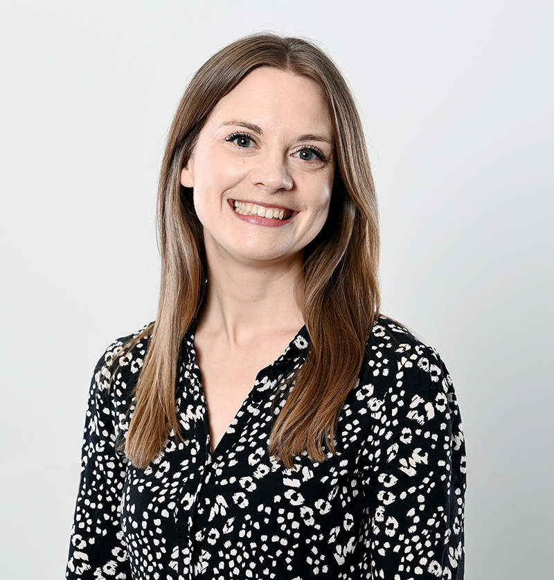 Caroline Julian, with straight, light brown hair, is smiling at the camera. She is wearing a black blouse with a white spotted pattern and stands against a plain, light grey background.