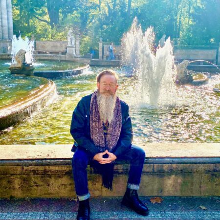 An older man with a long white beard, wearing a scarf, dark jacket, and boots, sits on a stone ledge in front of a large fountain in a sunlit park—perhaps reflecting on his role in the Community Advisory Group. Trees and statues are visible in the background.