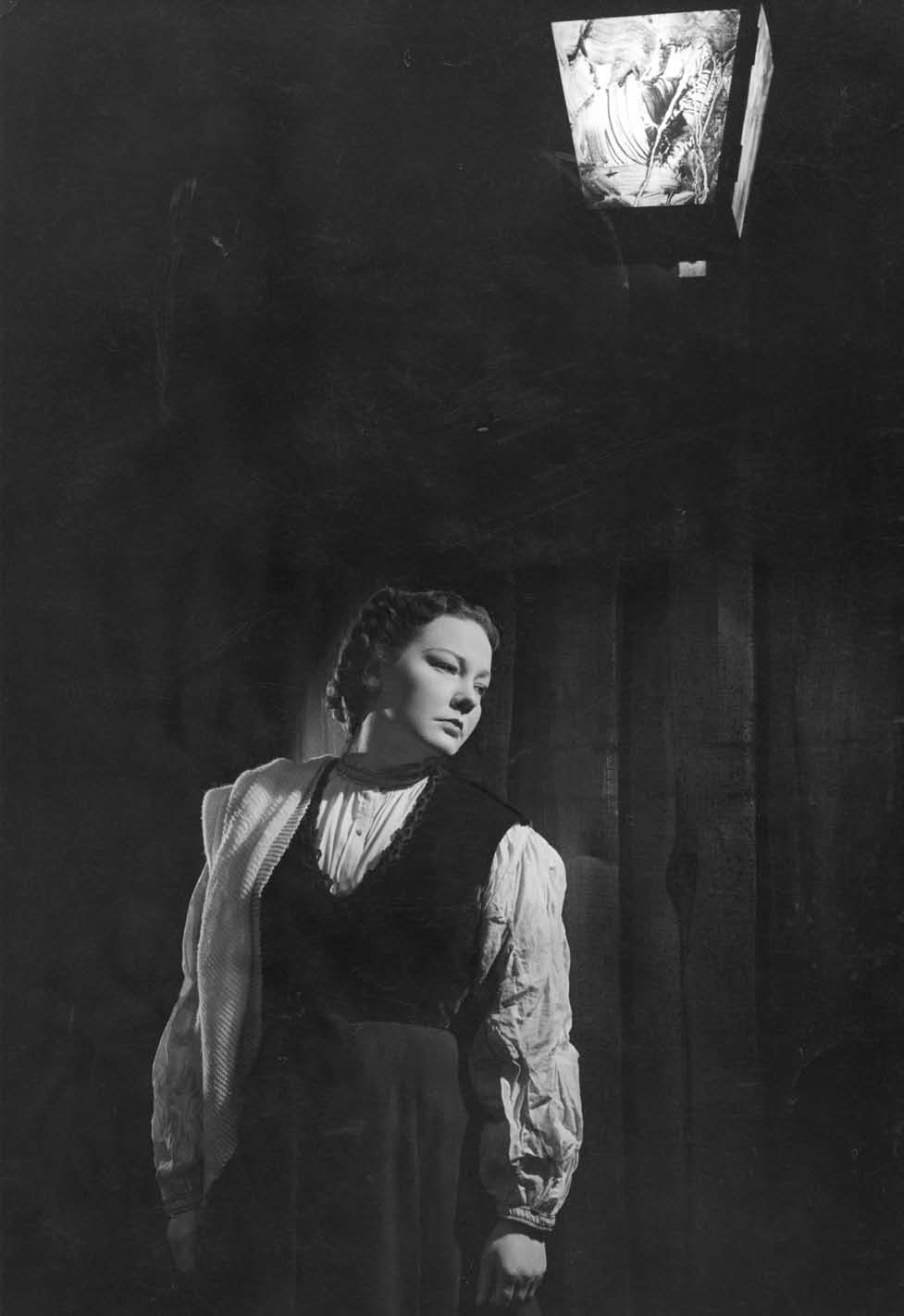 A woman in vintage clothing stands under a lit lantern, looking down thoughtfully. The dark background highlights her face and the illuminated lamp above her.