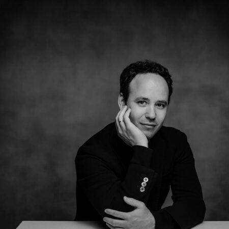 Gergely Madaras, a man with short dark hair, sits at a table in a dark jacket with visible buttons, resting his head on his hand and looking at the camera. The black and white photo features a plain background.