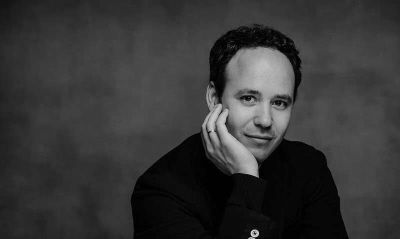 Gergely Madaras, a man with short dark hair, sits at a table in a dark jacket with visible buttons, resting his head on his hand and looking at the camera. The black and white photo features a plain background.
