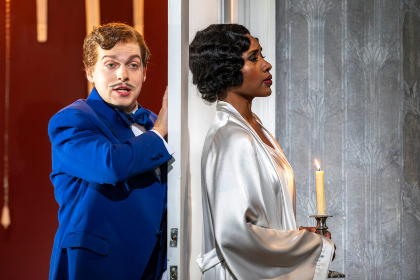 A man in a blue dinner jacket stands on one side of a doorway, looking and smiling towards a woman on the other side. The woman, wearing a white satin dressing gown, holds a lit candle and gazes ahead with a serious expression.