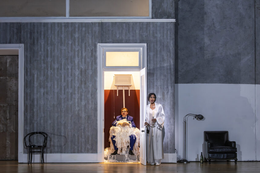 A person in a blue suit sits in a small room filled with white streamers, whilst another person in a white dressing gown stands outside the doorway holding a glass. The scene is set in a sparsely furnished, modern interior.