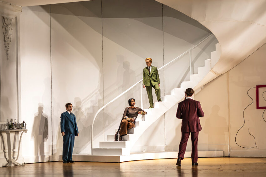Four people in formal attire stand and sit on a white, curved staircase and stage set with minimalist white walls, modern art, and sparse furniture, creating a dramatic theatrical scene.