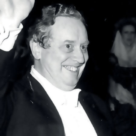 Charles Mackerras, in formal attire, smiling broadly and raising one arm in a wave. The background is dark, with indistinct figures visible.