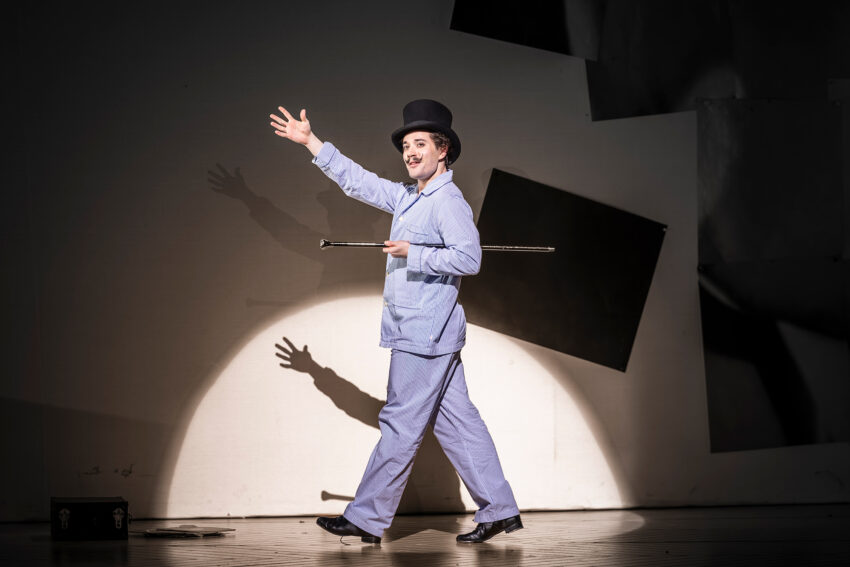 A performer dressed in light blue clothes, a bowler hat, and holding a cane walks across a stage, smiling and waving, illuminated by a spotlight. Dark geometric shapes and shadows are visible on the background wall.