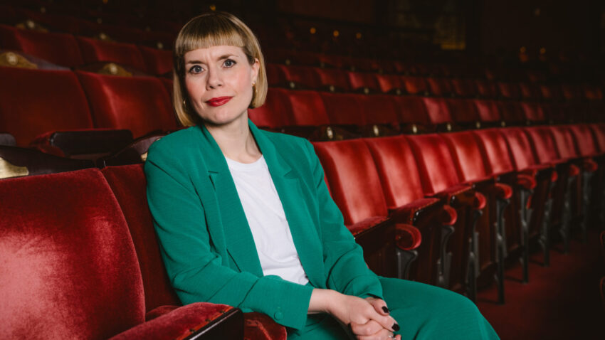 A woman with short blonde hair and red lipstick, wearing a green suit and white shirt, sits alone in a row of red theatre seats, looking at the camera.