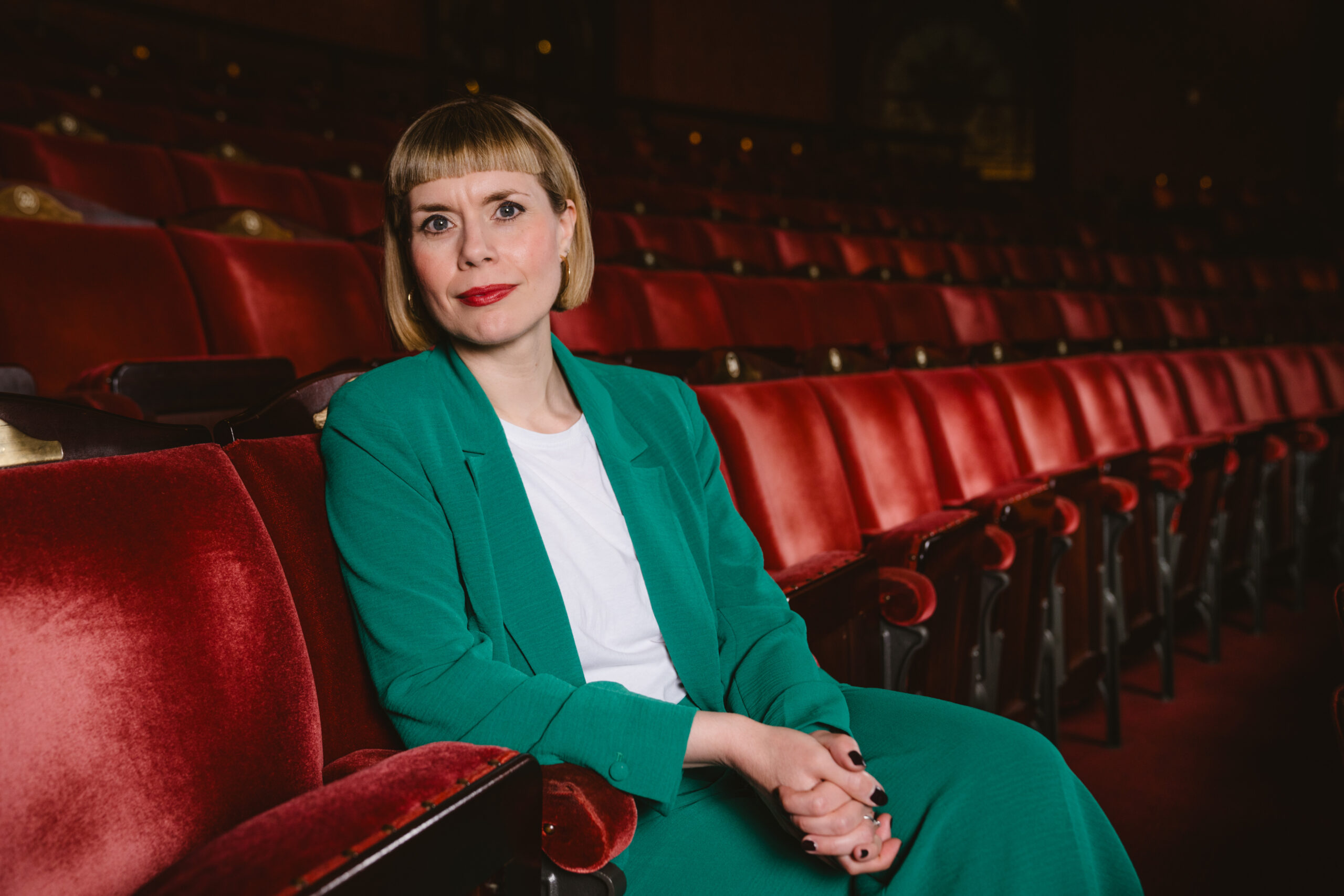 A woman with short blonde hair and red lipstick, wearing a green suit and white shirt, sits alone in a row of red theatre seats, looking at the camera.