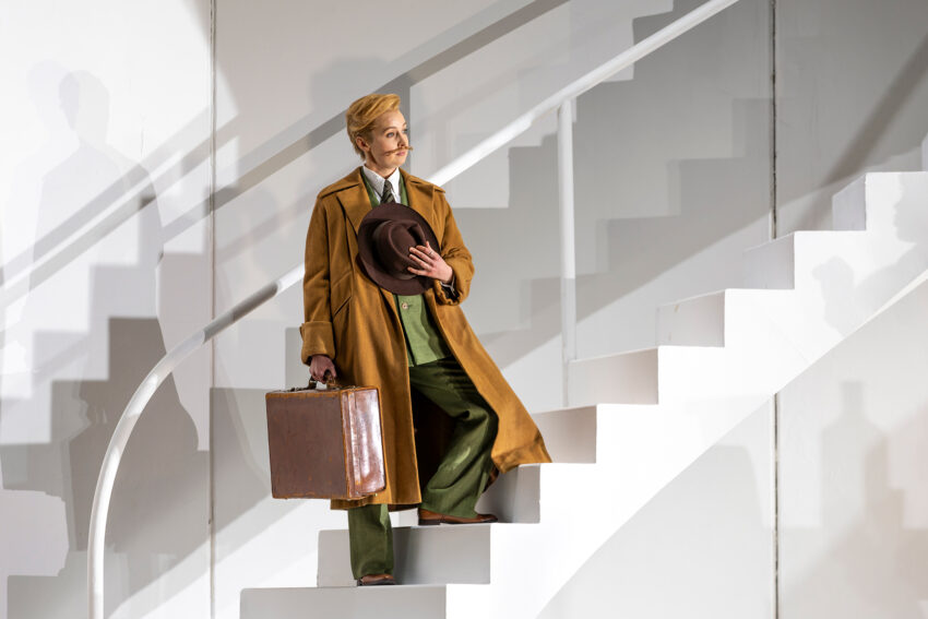 A person in a long brown coat holds a hat and a vintage suitcase while standing on a white, curved staircase in a brightly lit, minimalist setting.