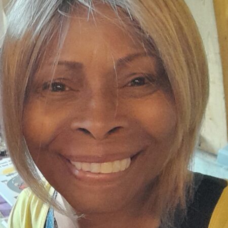 A woman with straight, shoulder-length blonde hair smiles warmly at the camera. She is wearing a yellow top with a dark lace trim underneath, ready for a Community Advisory Group meeting. The softly lit background features wooden elements and some papers.