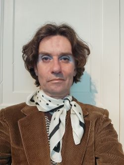 A person with wavy brown hair, member of the Community Advisory Group, wears blue eyeshadow, a white scarf with black dots, and a brown corduroy jacket, standing in front of a white panelled door.