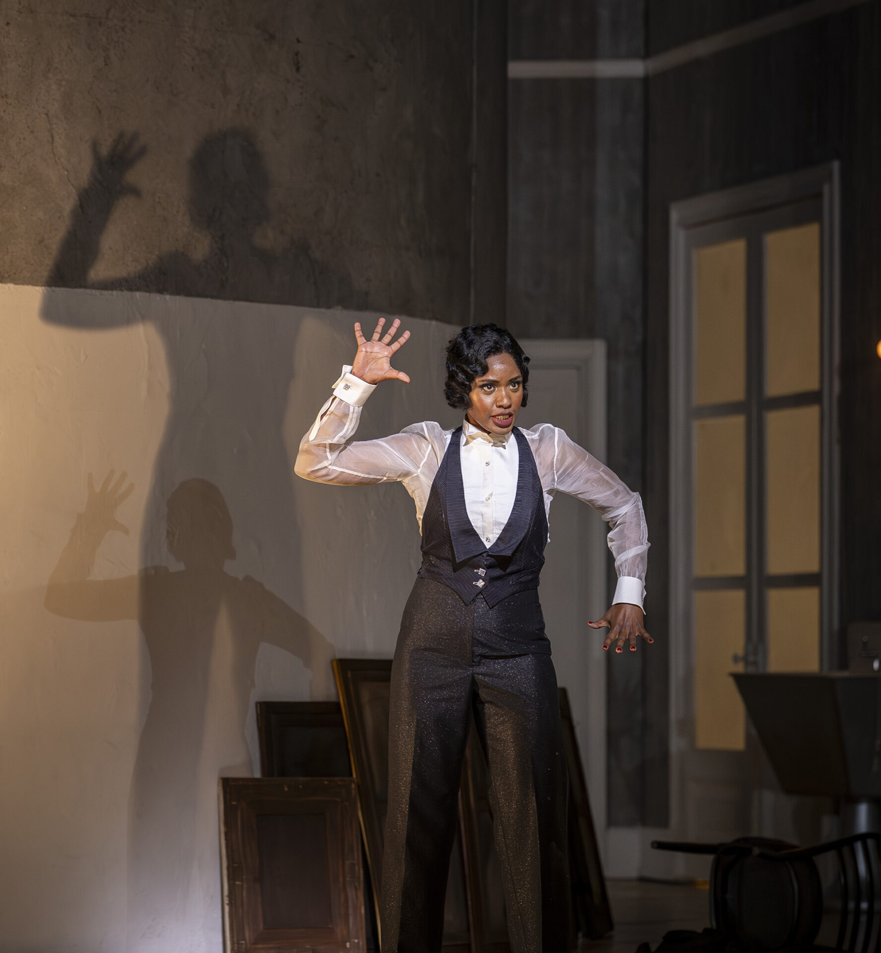 A person in a tuxedo and sheer white shirt stands on a stage with one hand raised, casting a shadow on the wall behind them. The background includes empty picture frames and an upturned chair.