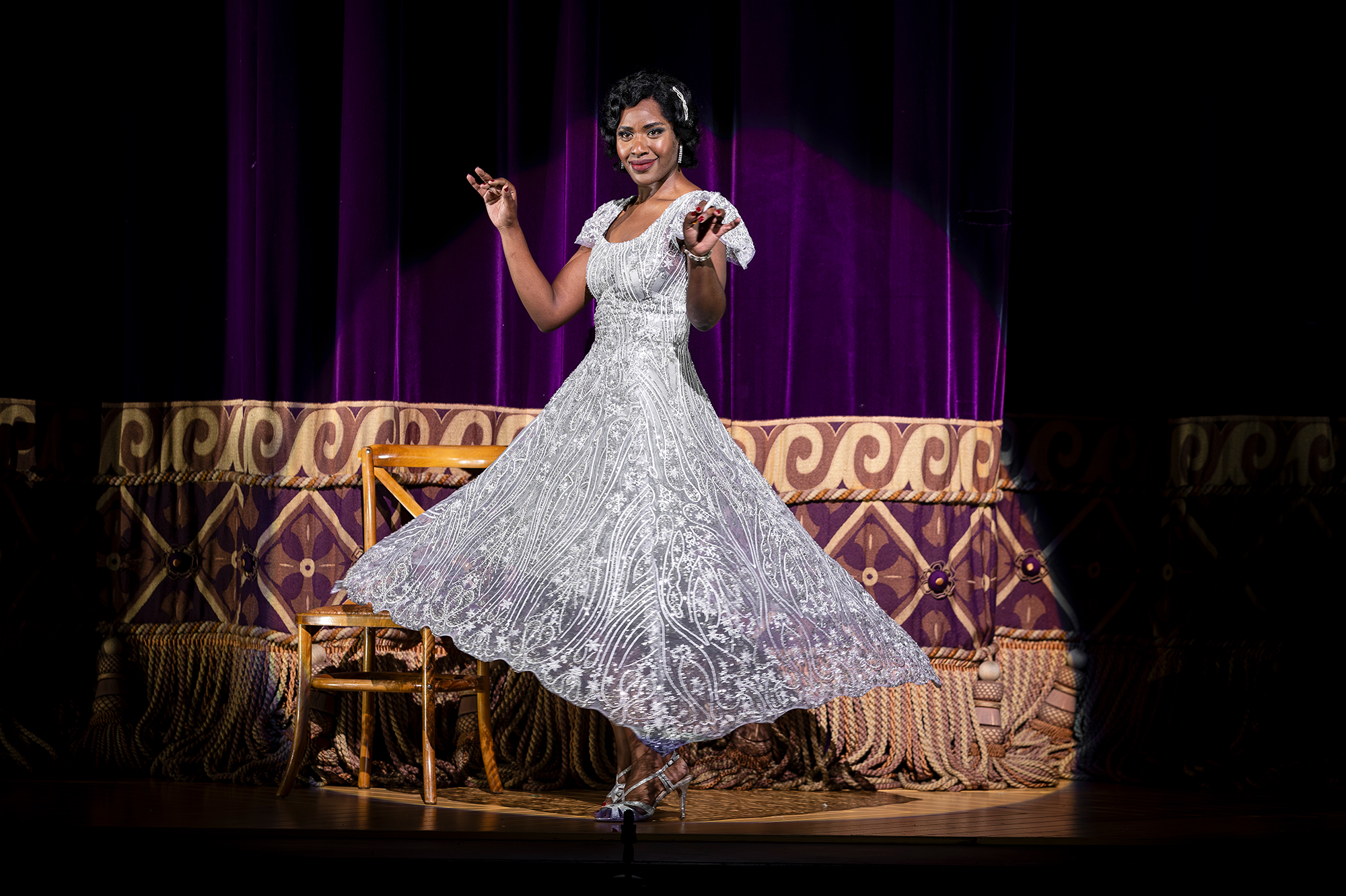 A woman in a sparkling white dress poses gracefully on stage in front of a purple curtain, with a wooden chair behind her. The dress fans out elegantly as she lifts her arms and smiles.