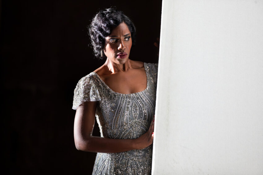 A woman in an elegant, glittering dress stands partly in shadow, looking to the side with a serious expression, one hand resting on a large white panel.