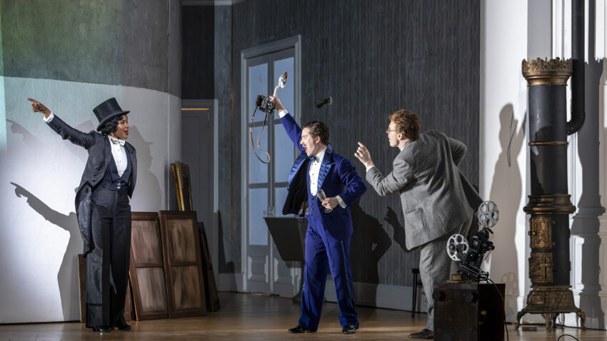 Three actors on a stage perform dramatically; one in a dinner jacket points left, another in a blue suit holds a camera, and a third in glasses gestures beside an old film projector and ornate column.