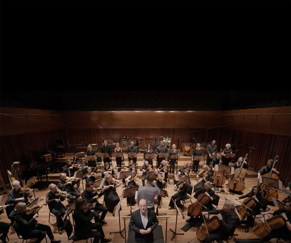 Orchestra of ENO sits on stage in a concert hall, with musicians playing string and percussion instruments; the conductor, Matthew Kofi Waldron, in front of whom stands in front singer Neal Davies, facing the camera.
